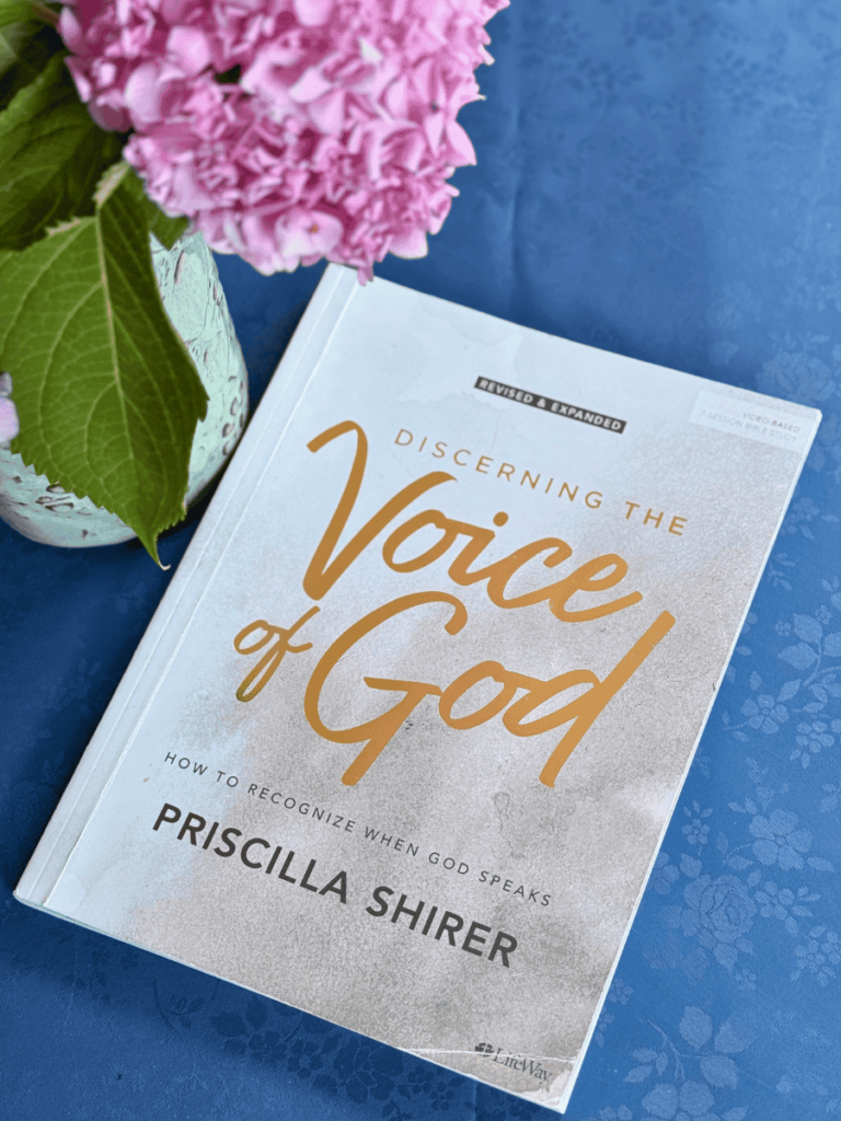 devotional "Discerning the Voice of God" by Priscilla Shirer laying on a table with a blue floral tablecloth next to a vase of pink hydrangeas