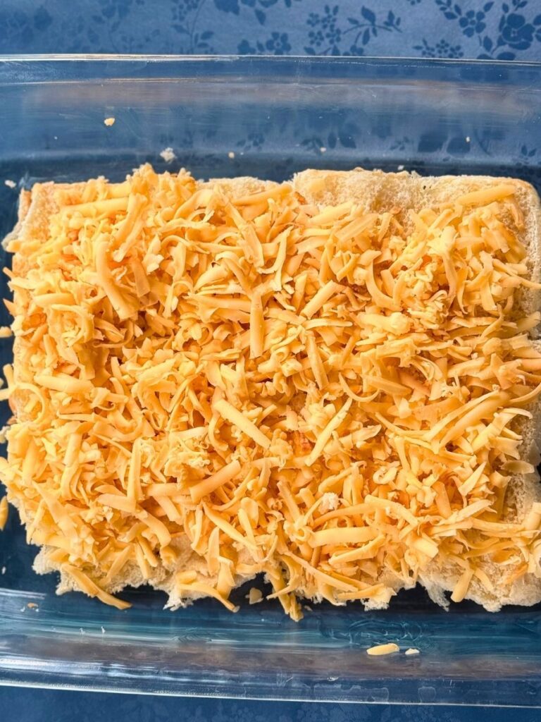 bottom half of hawaiian rolls topped with shredded gouda resting in a buttered 13" x 9" glass pan