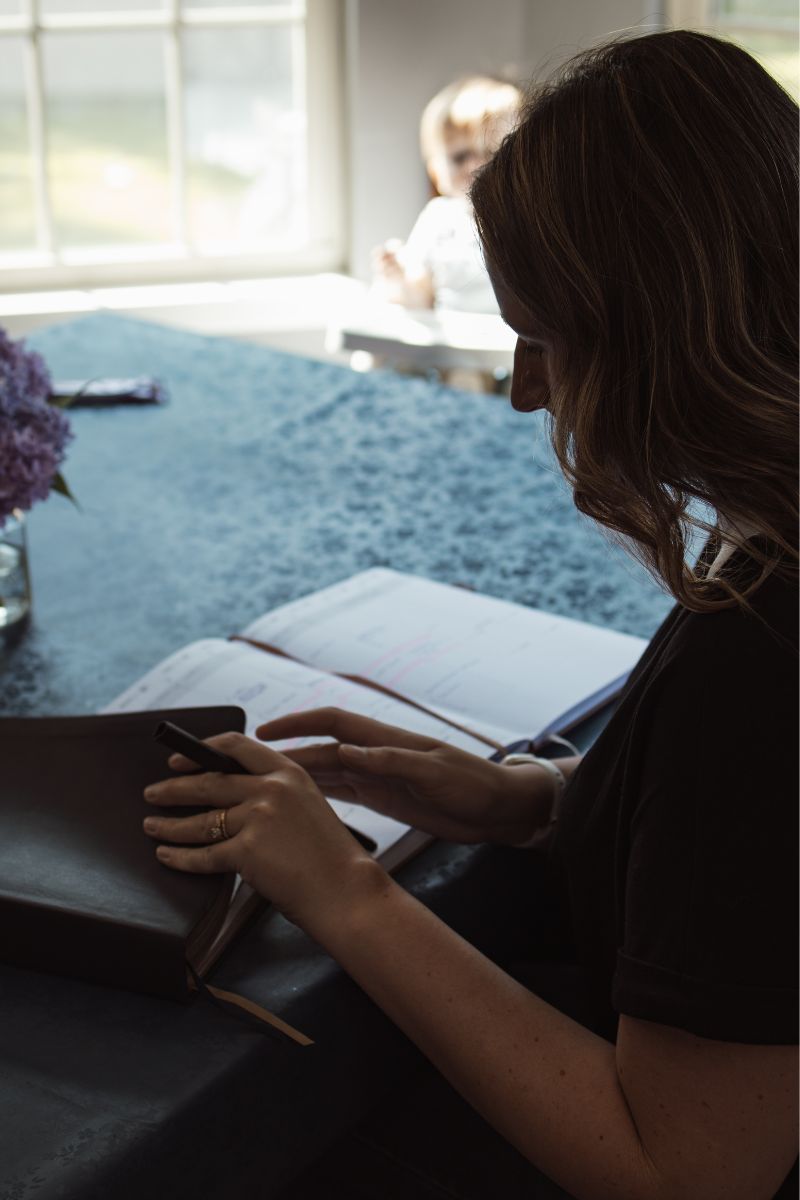 mom sitting at table looking at her opened planner and opening her bible while a baby sits in a high chair in the background